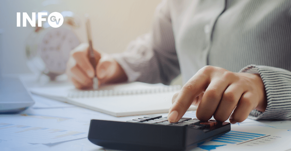 Mão de pessoa a calcular despesas com uma calculadora e a escrever num bloco de notas, com gráficos financeiros ao fundo
